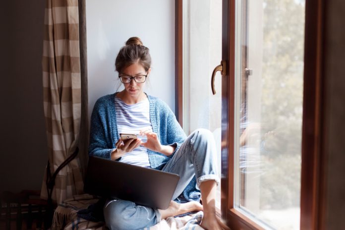 home office dziewczyna pracująca na laptopie i telefonie siedząca na oknie